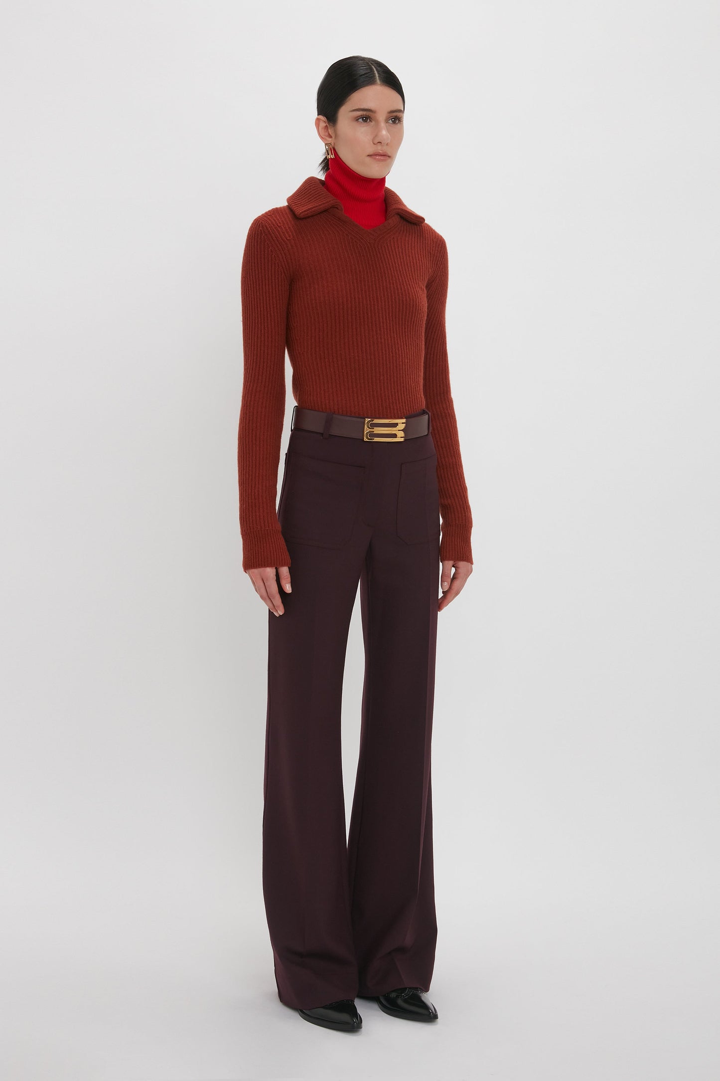 A person stands against a plain white background, wearing a red turtleneck, dark red sweater, and high-waisted Alina Trouser In Deep Mahogany from Victoria Beckham with a wide belt. The pants, made from recycled wool, feature a super-high waist. They have dark hair tied back and wear black shoes.