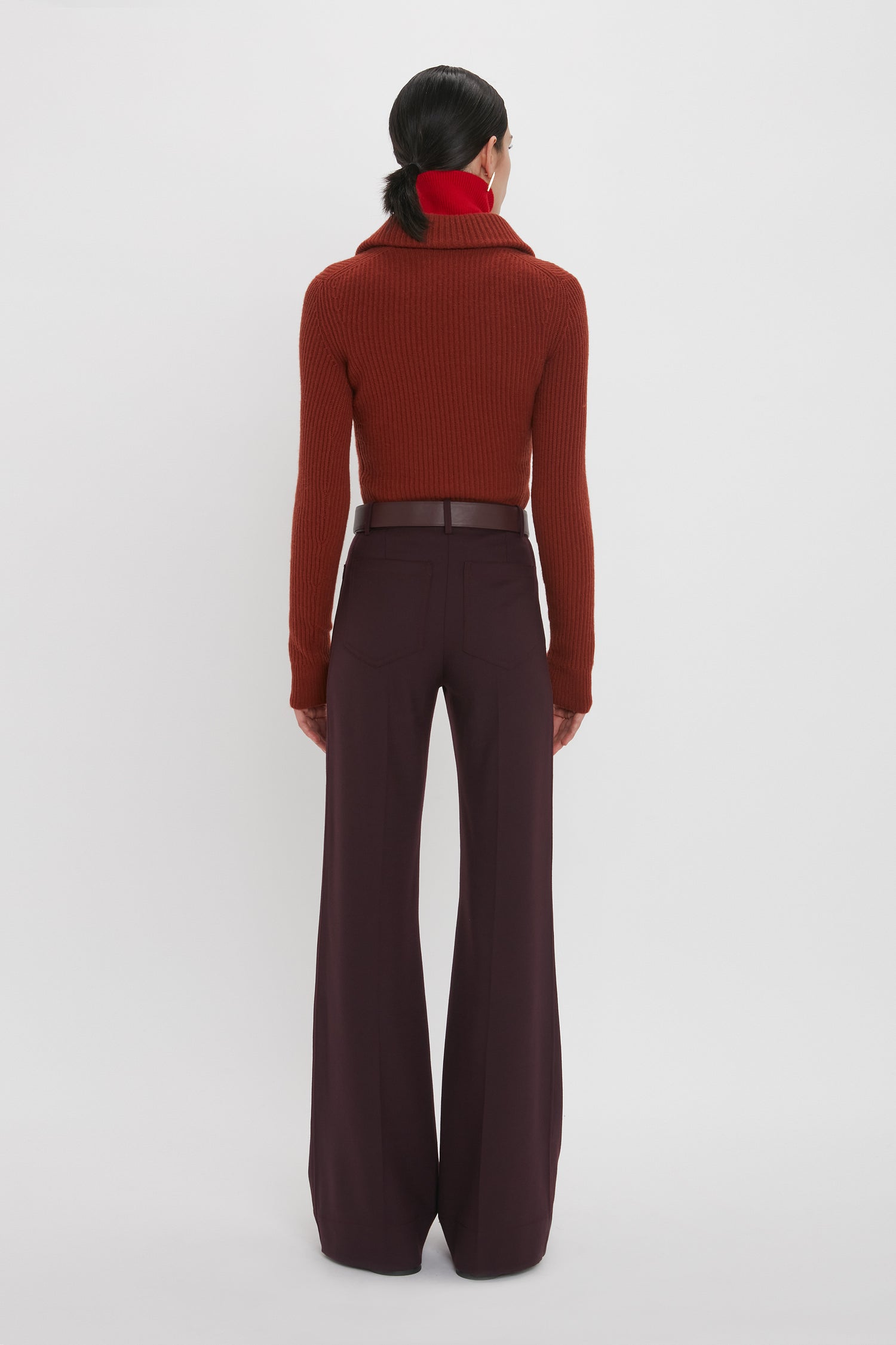 Back view of a person wearing a tactile Double Collared Jumper In Russet by Victoria Beckham and dark wide-leg pants, with hair tied back, standing against a plain white background.