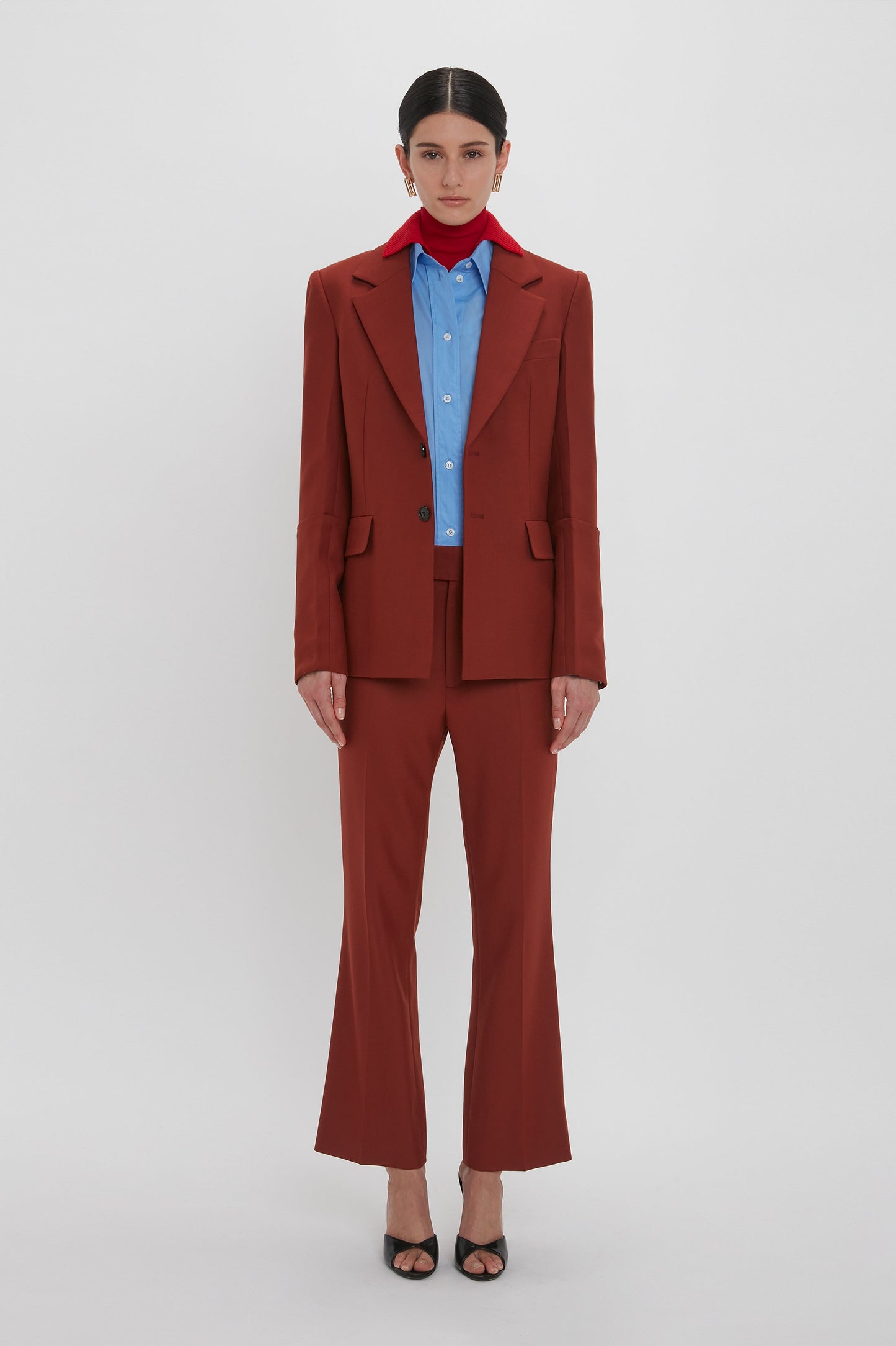 A person stands against a plain white background, wearing a rust-colored suit over a light blue button-up shirt and Wide Cropped Flare Trouser In Russet by Victoria Beckham, complemented by a red scarf around the neck and black open-toe heels.