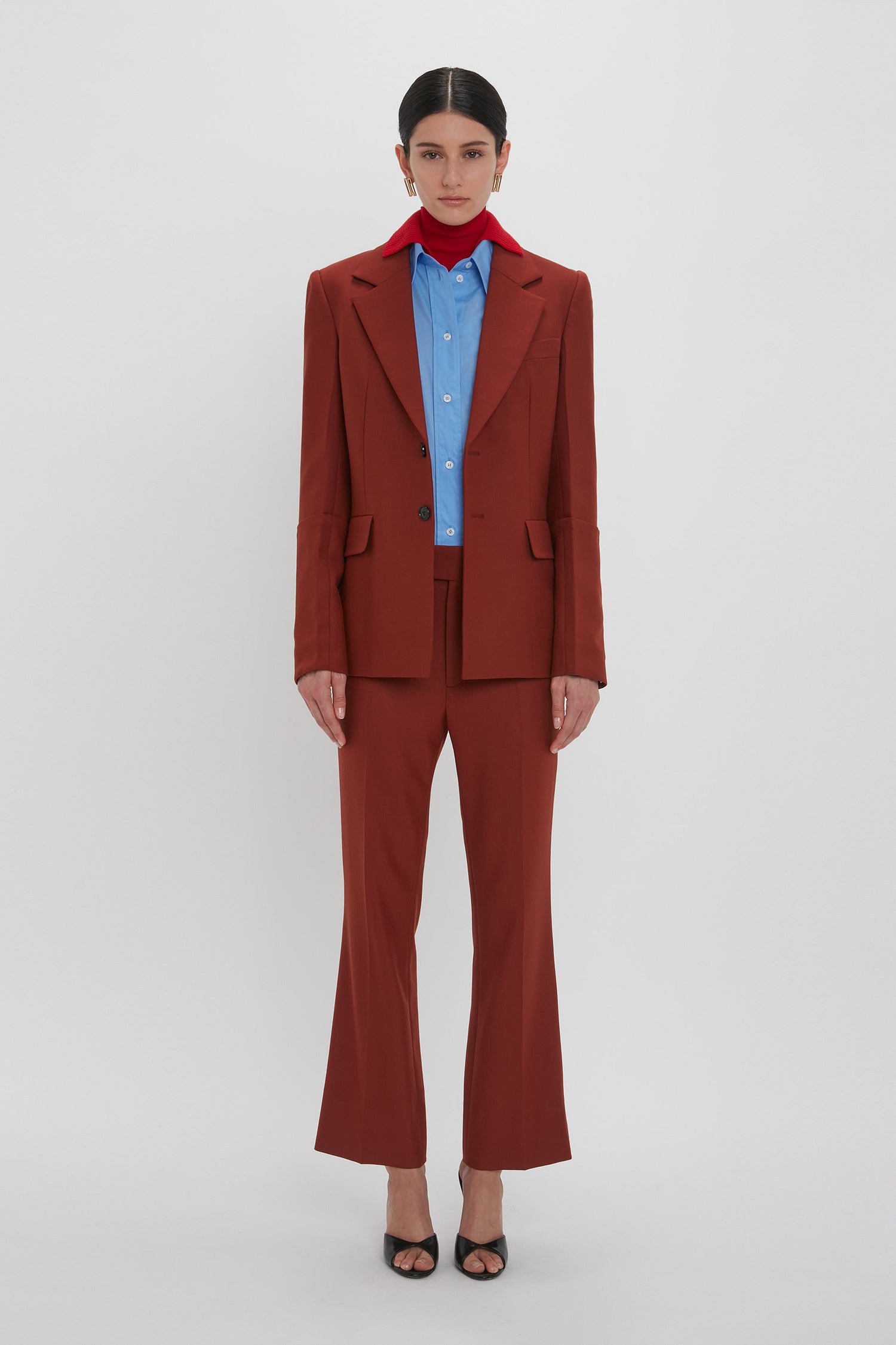 A person stands against a plain background, wearing the Sleeve Detail Patch Pocket Jacket In Russet from Victoria Beckham, paired with a blue shirt and red neck scarf. They are also wearing black open-toe heels.