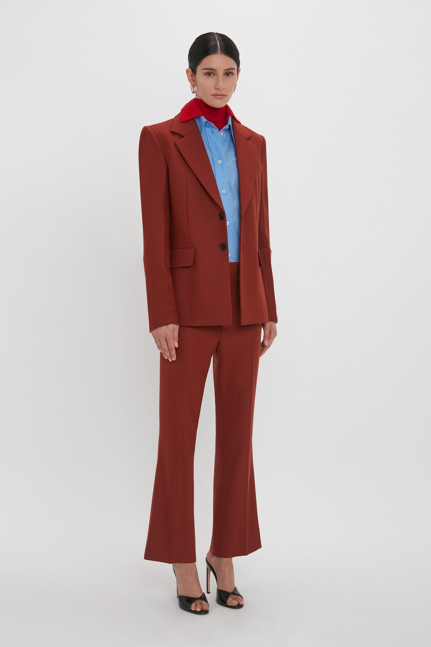 A person stands against a plain background wearing a rust-colored recycled wool suit, a blue shirt, red scarf, and black high-heeled shoes. The Victoria Beckham Sleeve Detail Patch Pocket Jacket In Russet features contemporary detailing, adding a modern touch to the classic ensemble.