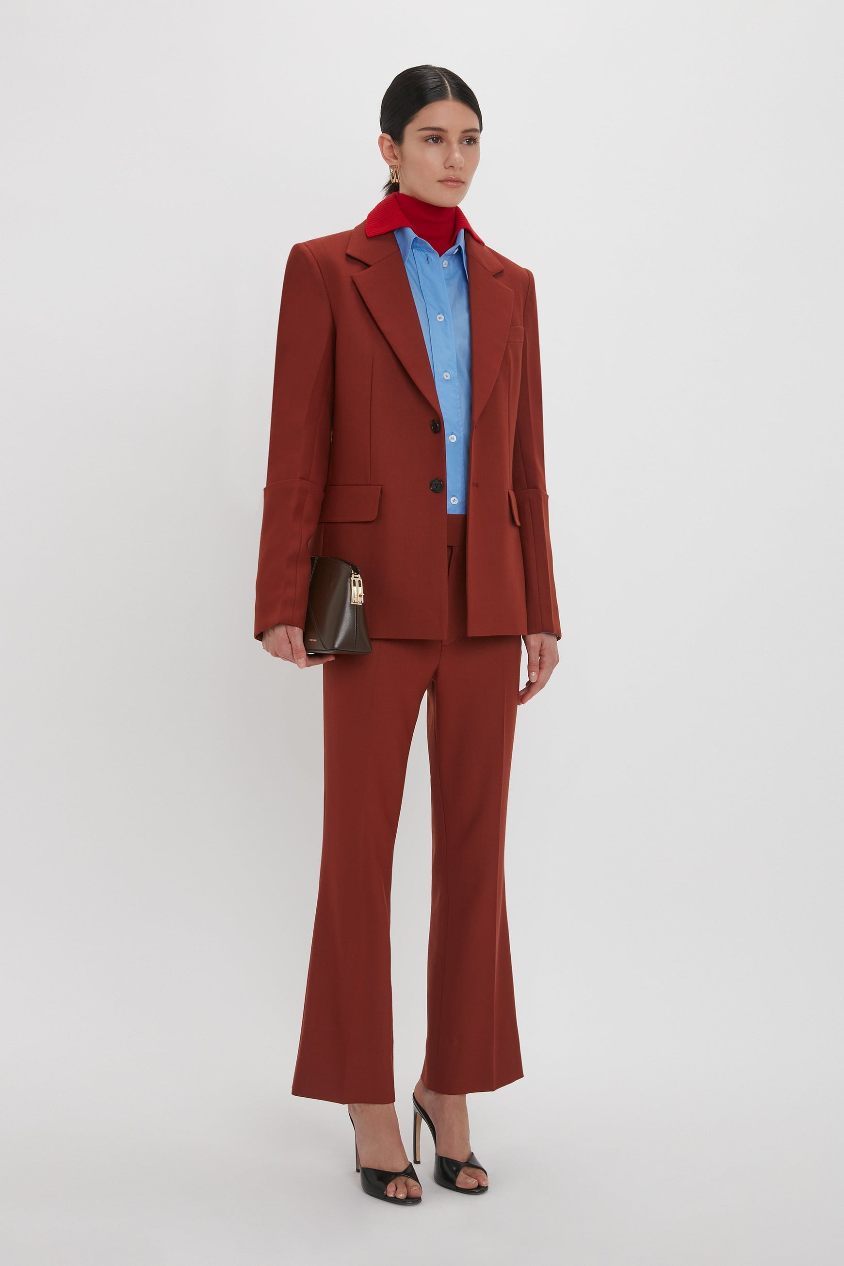 Person stands against a plain background wearing a red Sleeve Detail Patch Pocket Jacket In Russet by Victoria Beckham, a blue shirt, and black heels. They hold a brown handbag and have a red scarf tied around their neck, showcasing contemporary detailing.