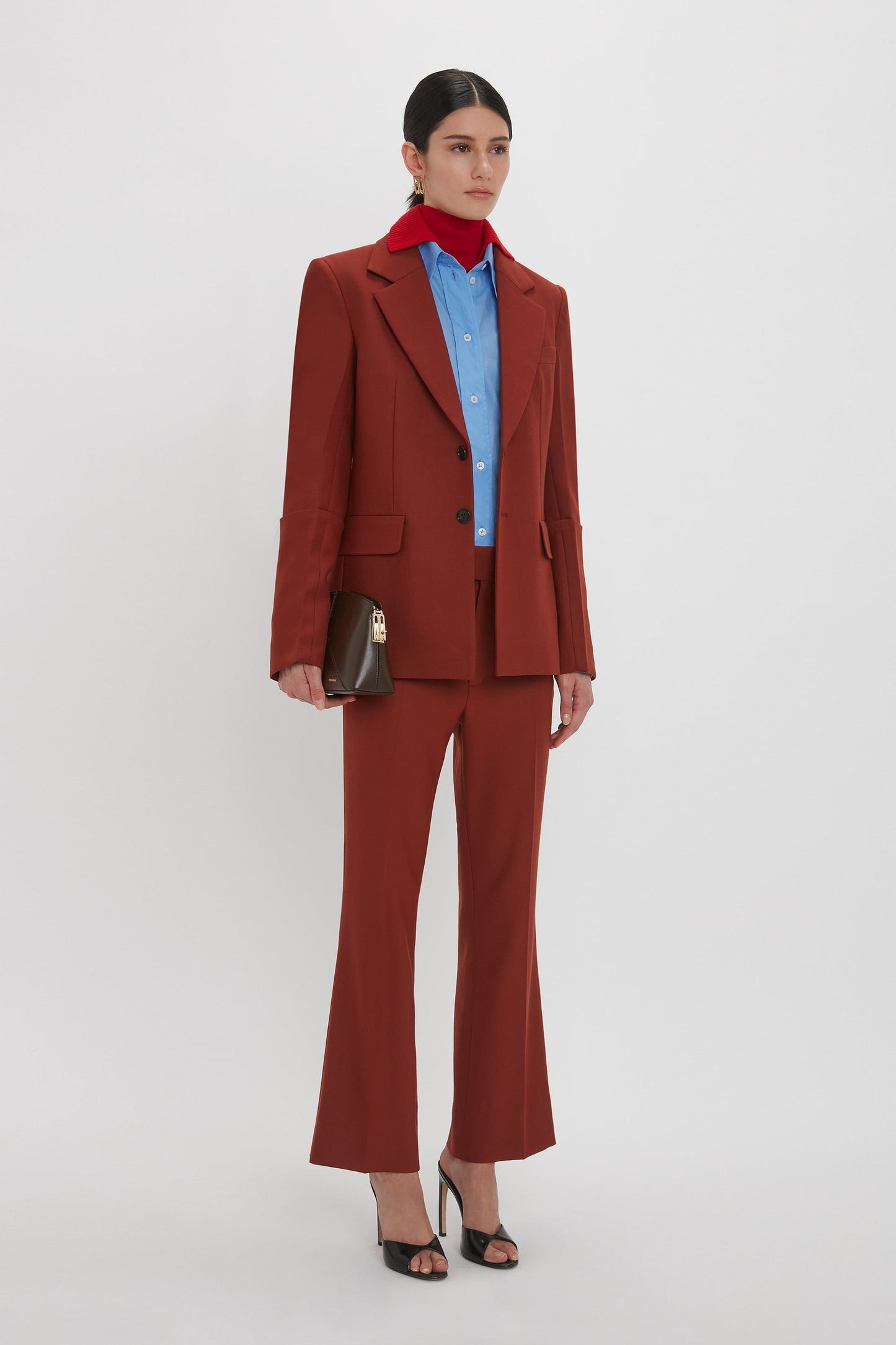 Person stands against a plain background wearing a red Sleeve Detail Patch Pocket Jacket In Russet by Victoria Beckham, a blue shirt, and black heels. They hold a brown handbag and have a red scarf tied around their neck, showcasing contemporary detailing.