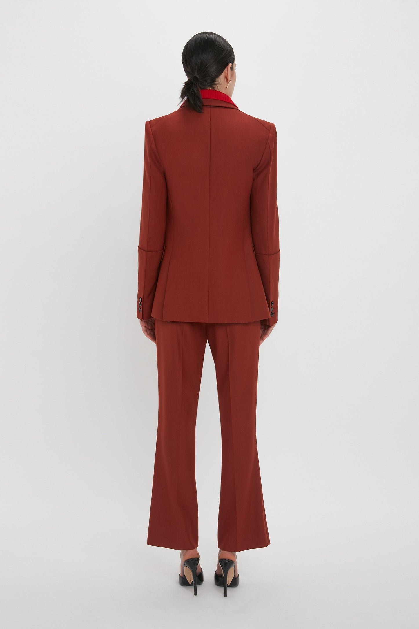 A person with black hair is shown from the back, wearing a red tailored suit with 1970s-inspired Wide Cropped Flare Trouser In Russet and black high-heeled shoes, standing against a plain white background—a nod to Victoria Beckham’s highlights for AW24.