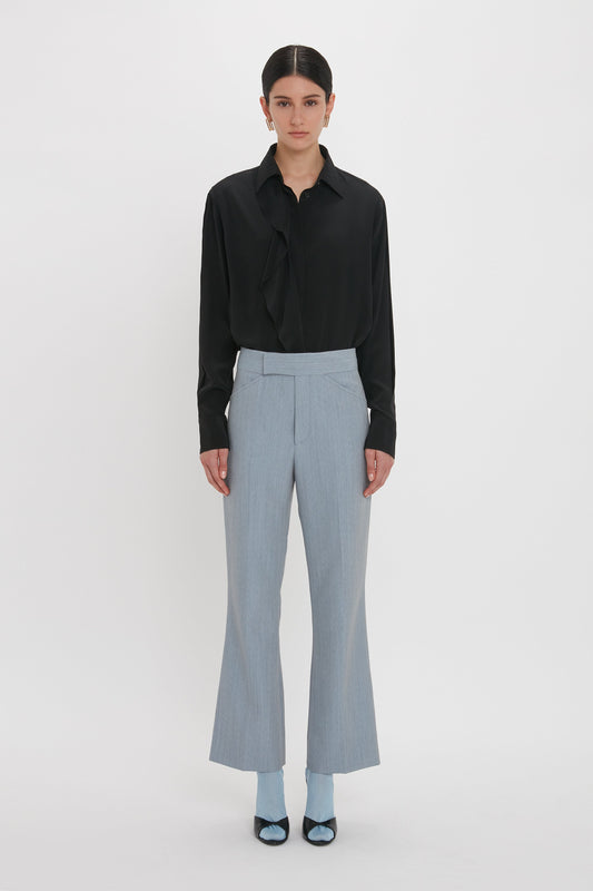 A person stands against a plain white background, wearing a black long-sleeve shirt, Exclusive Wide Cropped Flare Trouser In Marina by Victoria Beckham, blue socks, and black shoes.