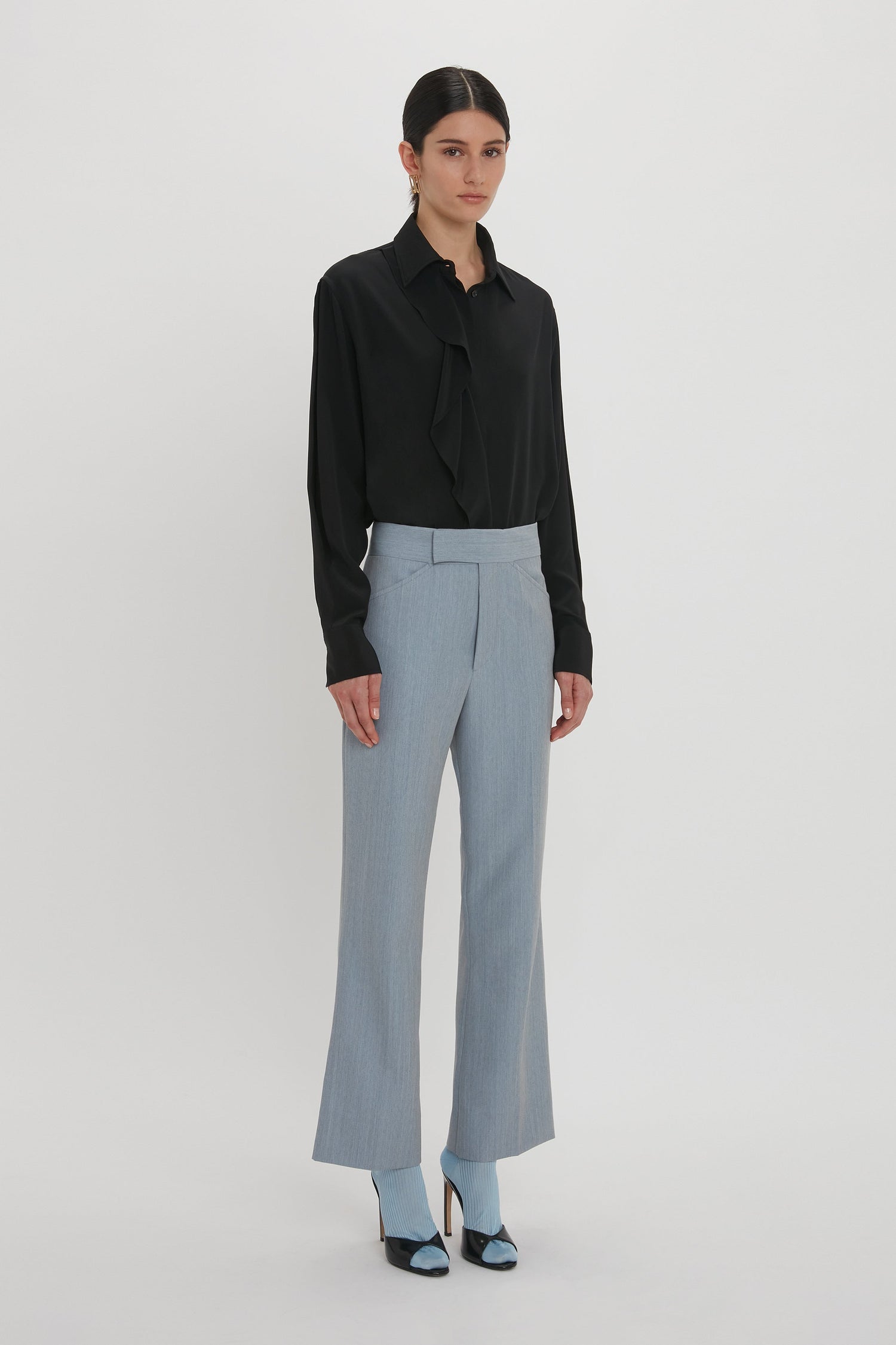 A person stands against a white background, wearing a black long-sleeve blouse, Victoria Beckham Exclusive Wide Cropped Flare Trouser In Marina, and light blue heels.