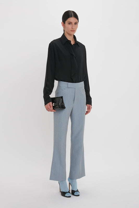 A person stands against a white background, wearing a black blouse, Victoria Beckham Exclusive Wide Cropped Flare Trouser In Marina, and black and blue heeled shoes. They are holding a black handbag.