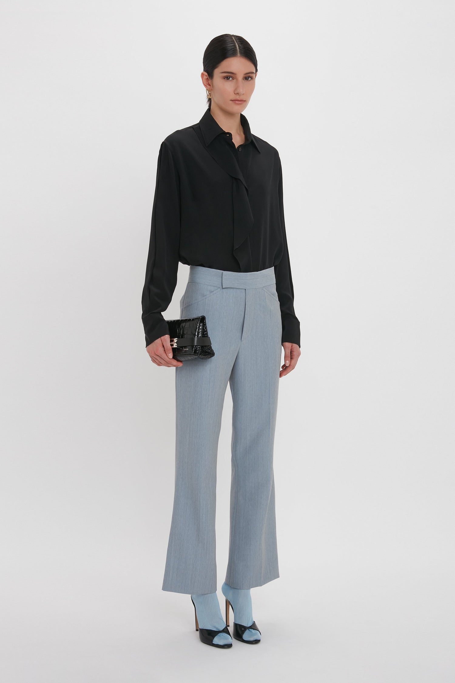 A person stands against a white background wearing a black blouse, light blue trousers, and black heels, holding a Mini B Pouch Bag In Croc Effect Black Leather by Victoria Beckham.