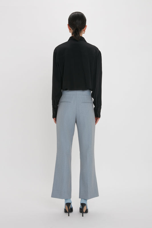 A person stands facing away from the camera, wearing a black blouse, Victoria Beckham Exclusive Wide Cropped Flare Trouser In Marina, and black high-heeled shoes.