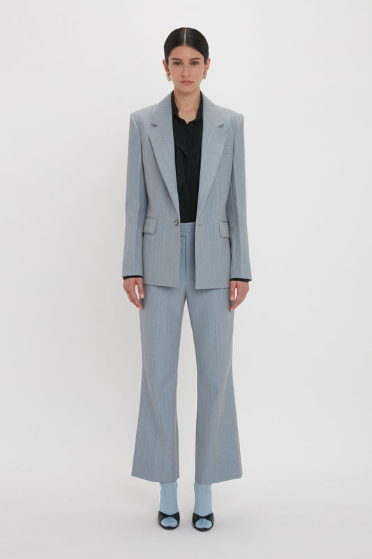Person standing against a plain background wearing an impeccably tailored light gray Exclusive Sleeve Detail Patch Pocket Jacket In Marina by Victoria Beckham and matching pants, paired with a black shirt and vibrant blue socks with black shoes.