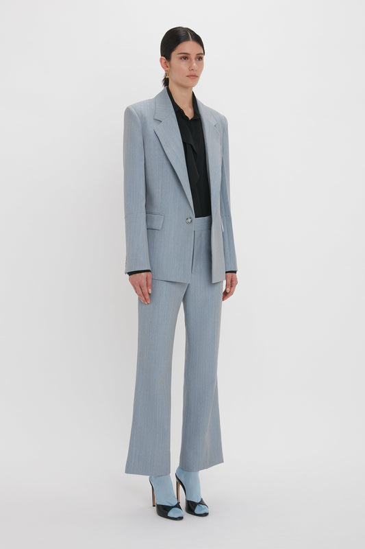 A person stands against a white background, wearing an impeccably tailored light blue pantsuit with the Exclusive Sleeve Detail Patch Pocket Jacket In Marina by Victoria Beckham, paired with a black blouse and blue open-toe heels.