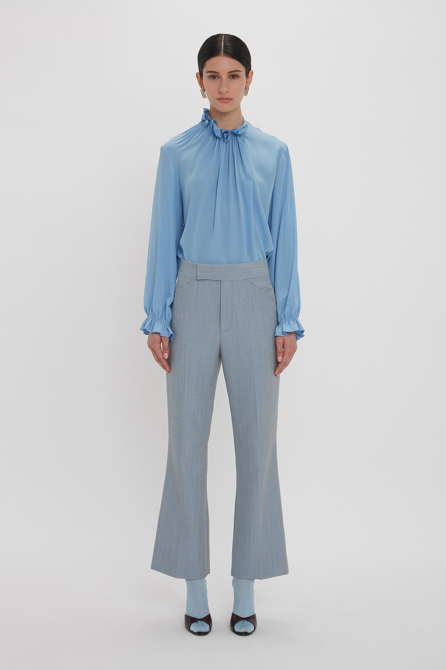 A person with dark hair is wearing a light blue Exclusive Ruffle Neck Blouse In Cornflower Blue by Victoria Beckham, light gray pants, and open-toe heels. They are standing against a plain white background.