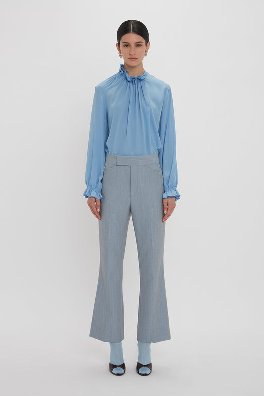 A person with dark hair is wearing a light blue Exclusive Ruffle Neck Blouse In Cornflower Blue by Victoria Beckham, light gray pants, and open-toe heels. They are standing against a plain white background.