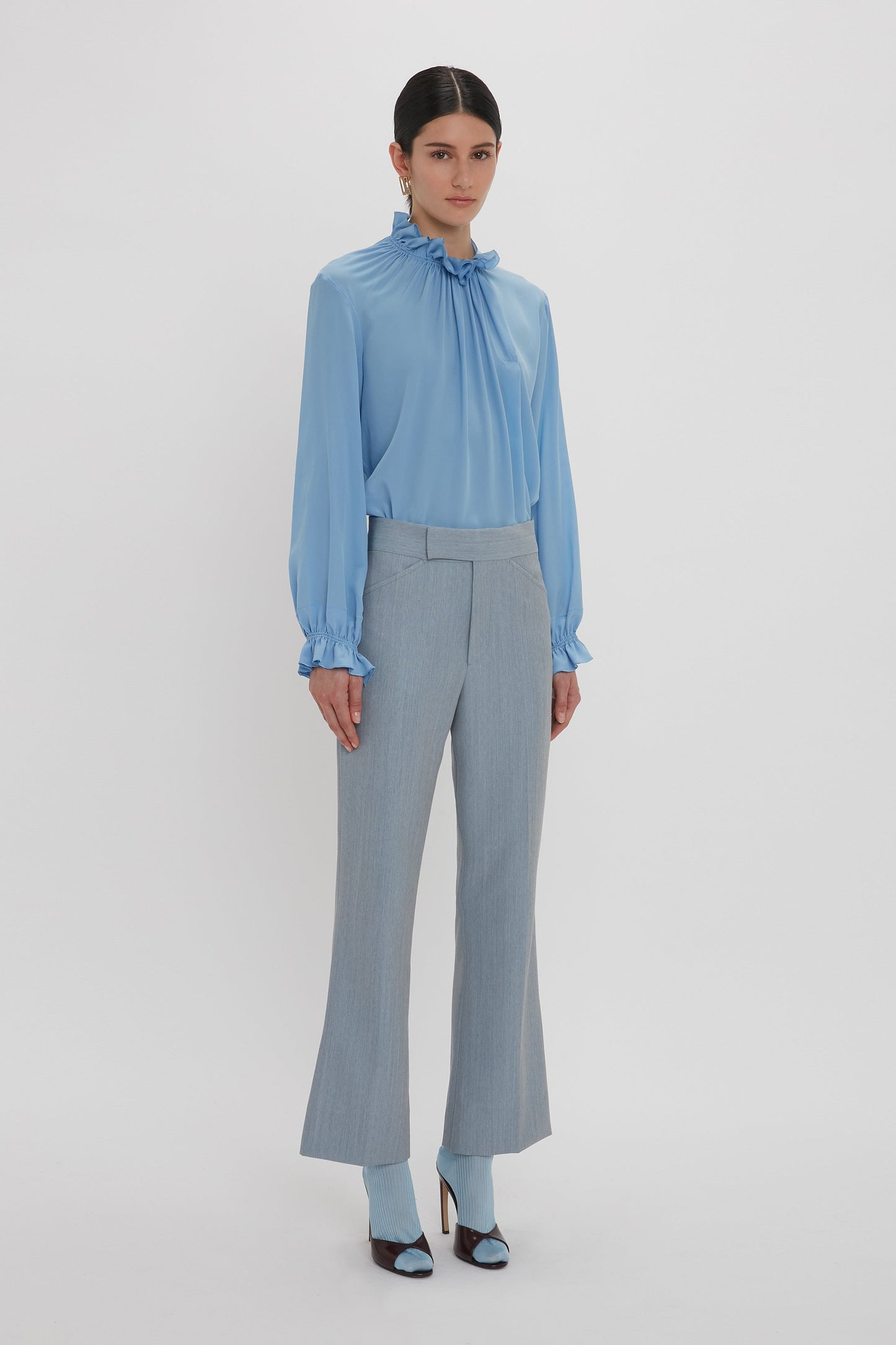 A person stands against a plain white background wearing the Exclusive Ruffle Neck Blouse In Cornflower Blue by Victoria Beckham, light grey pants, and blue high-heeled shoes.