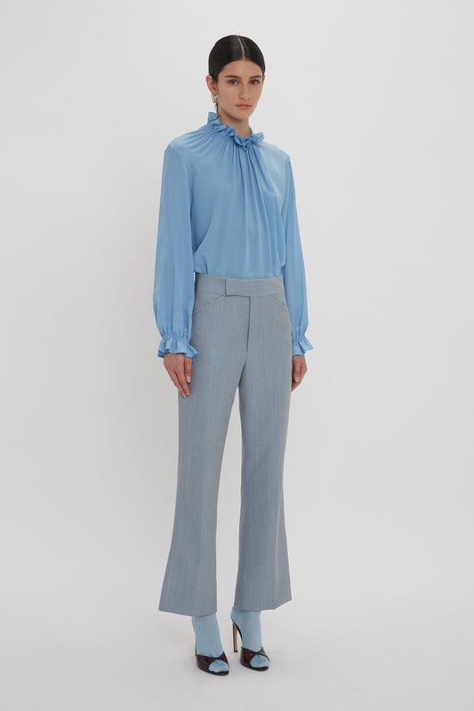 A person stands against a plain white background wearing the Exclusive Ruffle Neck Blouse In Cornflower Blue by Victoria Beckham, light grey pants, and blue high-heeled shoes.