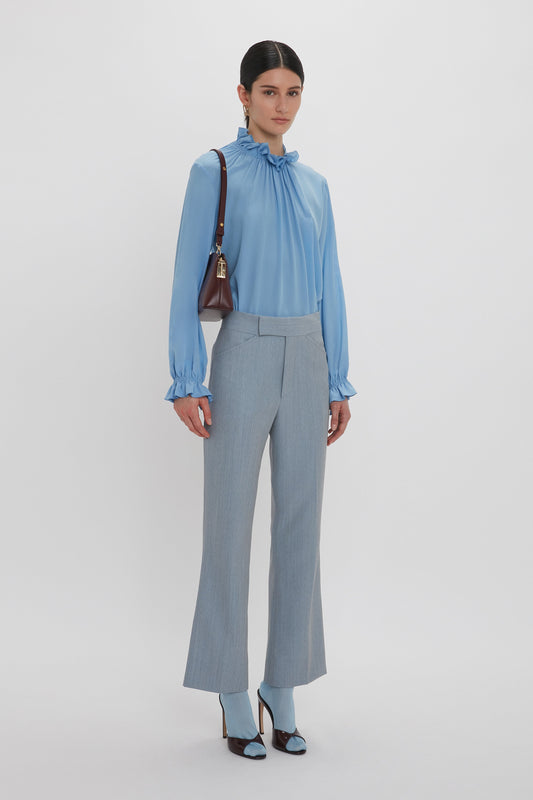 A person wearing a light gray, Exclusive Ruffle Neck Blouse In Cornflower Blue by Victoria Beckham, gray trousers, and blue high heels, holding a brown bag on their shoulder, stands against a plain white background.