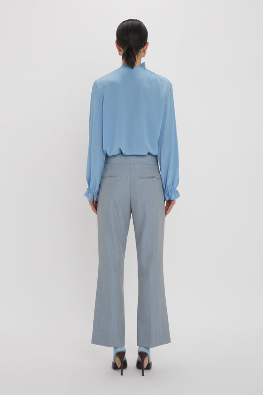 A person wearing a Victoria Beckham Exclusive Ruffle Neck Blouse In Cornflower Blue and grey pants stands facing away, showcasing the back view of their outfit.