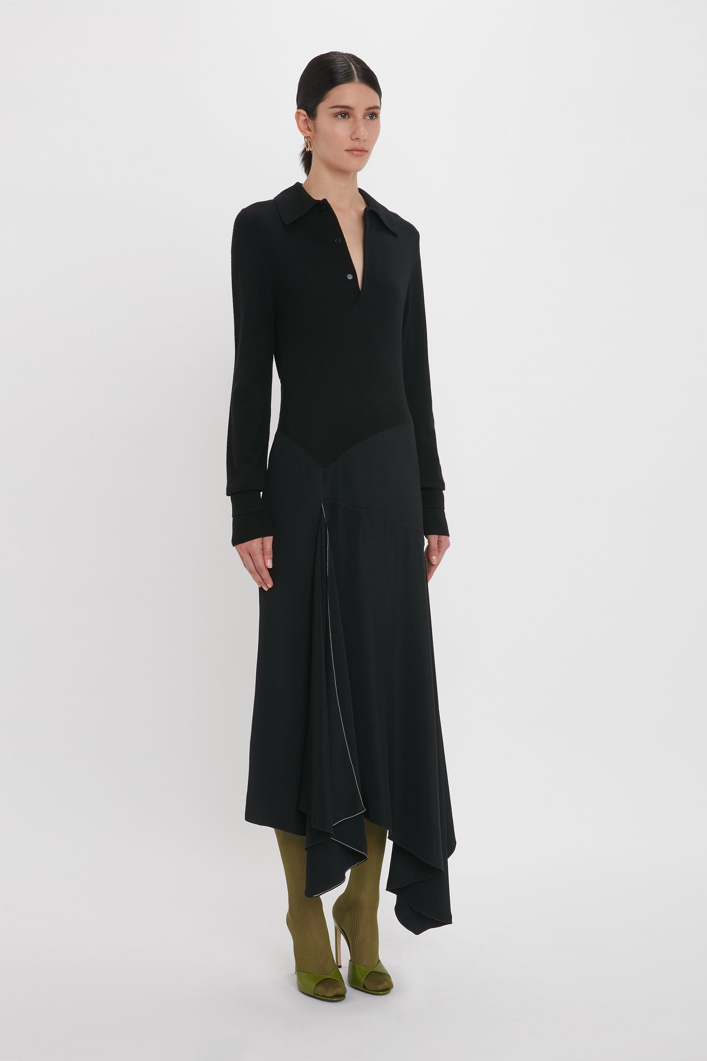 A person with dark hair is wearing a long-sleeved, black Victoria Beckham Henley Shirt Dress In Black with a V-neck and an asymmetrical hem, paired with olive green knee-high boots.