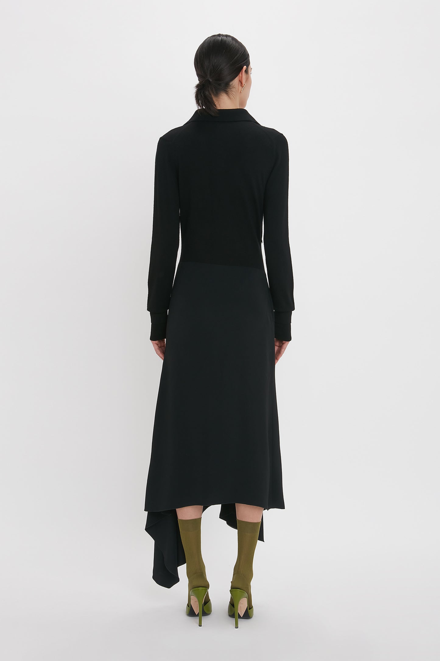Person standing, facing away, wearing a Henley Shirt Dress In Black by Victoria Beckham with an asymmetric waist seam and green high-heeled shoes against a plain white background.