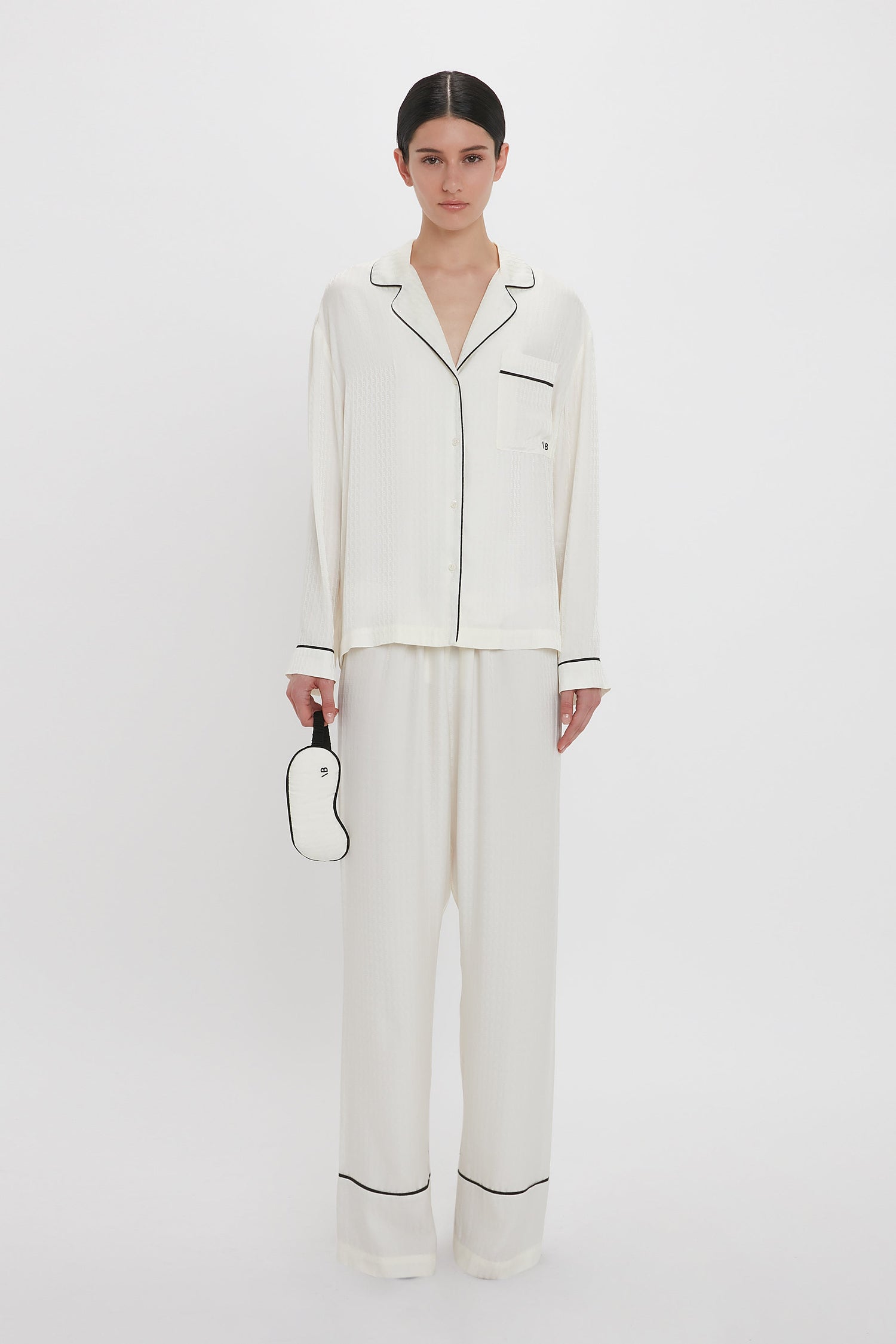 A person stands against a plain white background, wearing an elegant Victoria Beckham VB Monogram Pyjama Sleep Set In Ivory. They are holding a chic white sleep mask with a black outline in their right hand. The loungewear set exudes luxury, perfect for relaxing or sleeping comfortably.