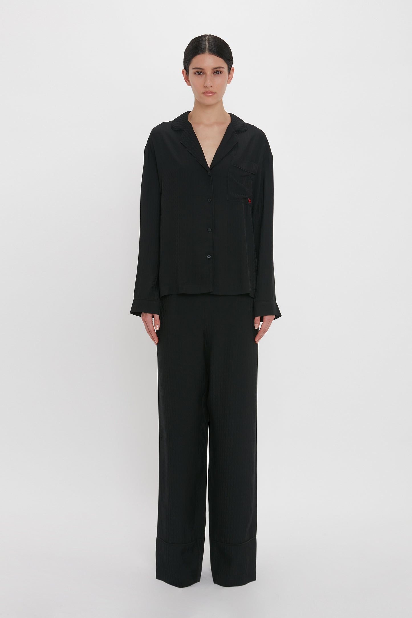 Person standing against a plain white background, dressed in the VB Monogram Pyjama Sleep Set In Black from Victoria Beckham.