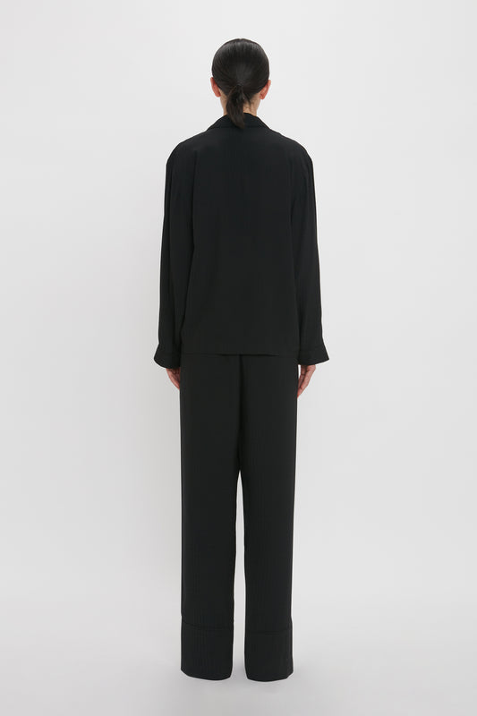 Back view of a person with dark hair tied back, standing against a plain white background, wearing a loose-fitting black long-sleeved top and black pants that resemble the VB Monogram Pyjama Sleep Set In Black by Victoria Beckham.