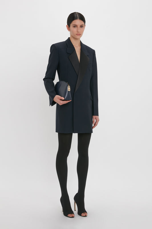 A person stands against a plain white background wearing a Victoria Beckham Exclusive Fold Shoulder Detail Dress In Midnight, black tights, and high heels, holding a small black purse.