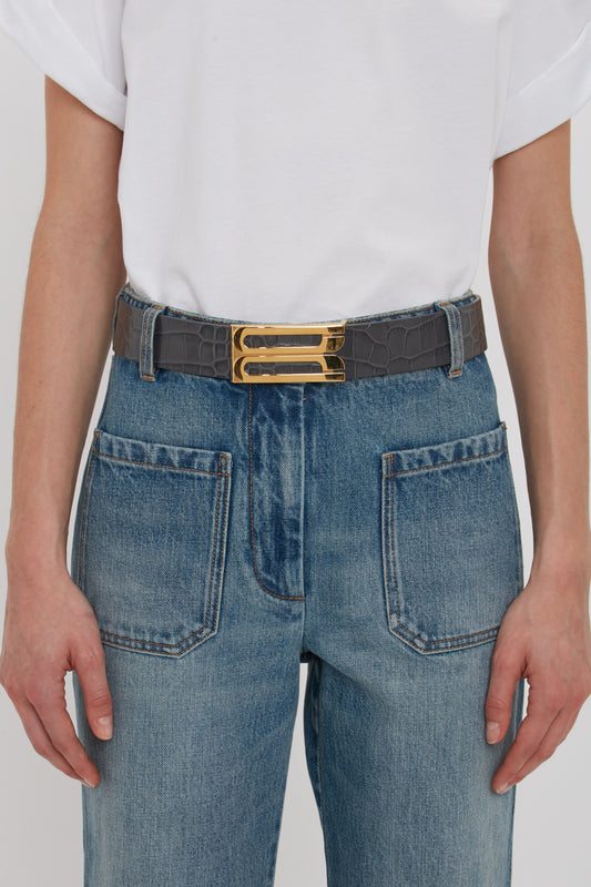A person dressed in a white shirt and blue jeans is shown wearing a Victoria Beckham Jumbo Frame Belt In Slate Grey Croc Embossed Calf Leather, featuring gold hardware.
