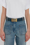 Person wearing a white t-shirt tucked into high-waisted blue jeans with a Jumbo Frame Belt In Midnight Blue Croc Embossed Calf Leather by Victoria Beckham featuring a large gold buckle.