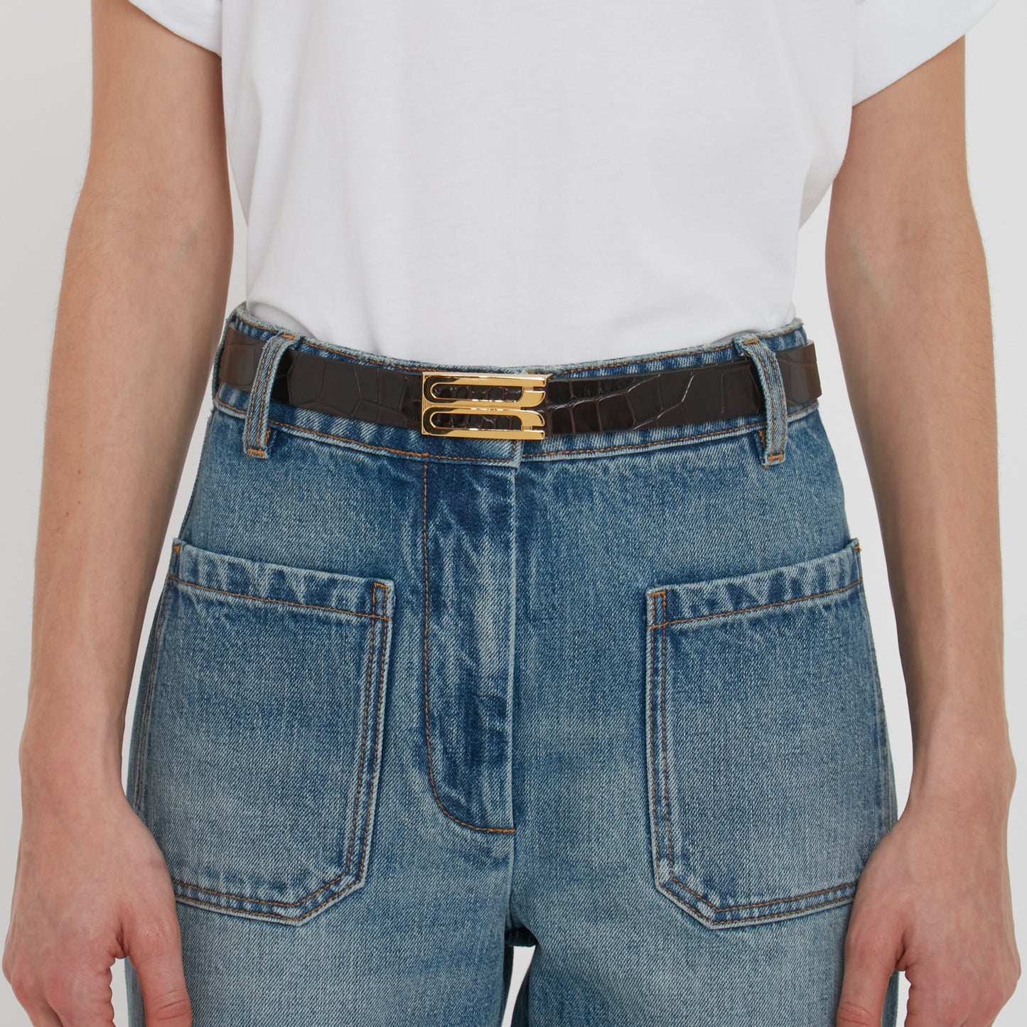 A person wearing a white shirt is shown from the waist down, dressed in high-waisted blue jeans with large front pockets and a Victoria Beckham Frame Belt In Espresso Croc Embossed Calf Leather adorned with gold hardware.