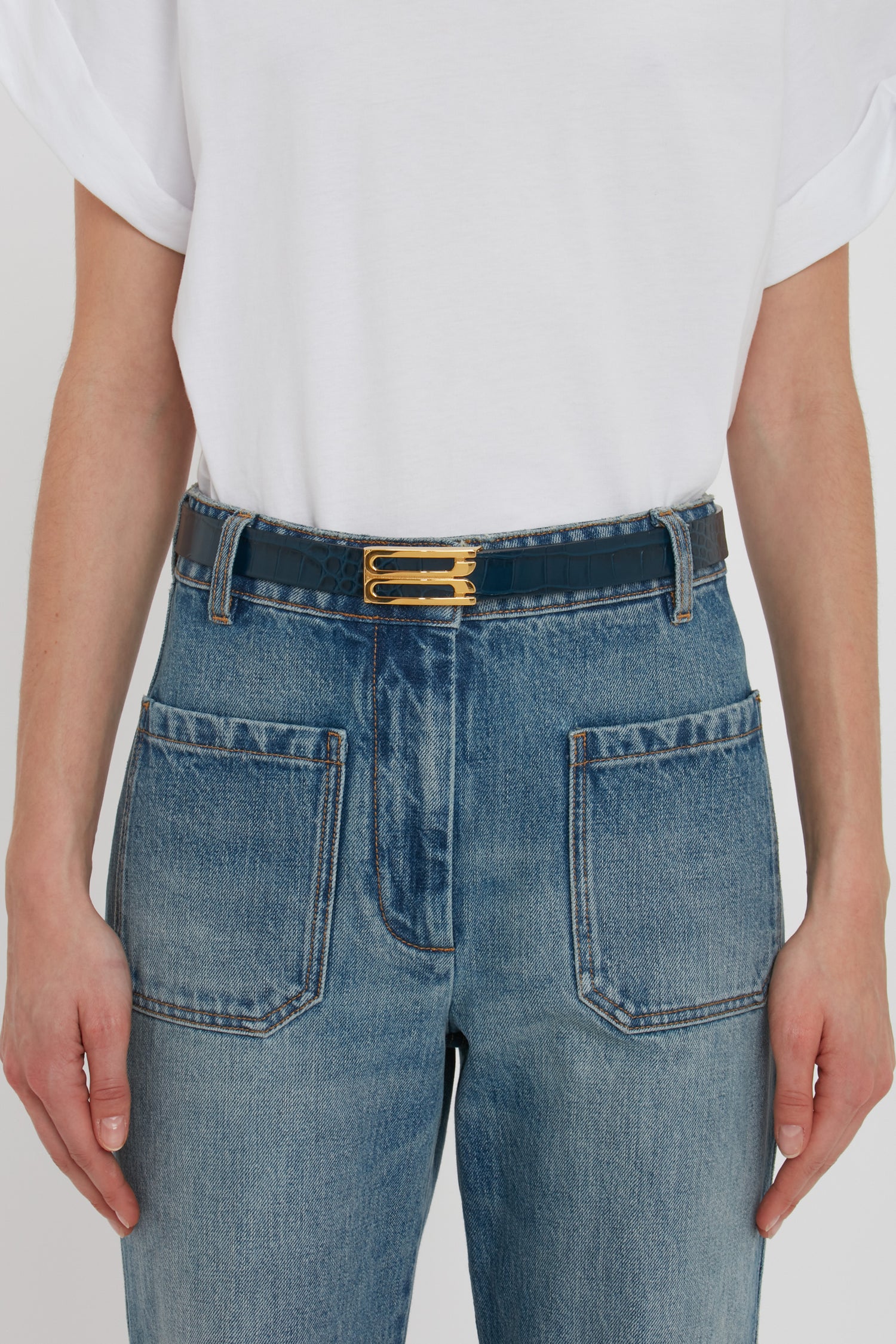 Person wearing a white t-shirt, blue jeans with large front pockets, and a Victoria Beckham Frame Belt In Midnight Blue Croc Embossed Calf Leather.