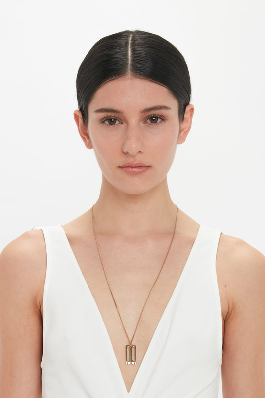A person with dark hair tied back wears a white V-neck top and a long necklace featuring the Victoria Beckham Exclusive Frame Necklace In Gold.