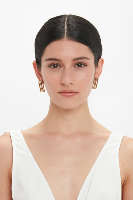A woman with dark hair pulled back, wearing Victoria Beckham Exclusive Frame Stud Earrings In Gold and a white sleeveless top, looks directly at the camera against a white background.