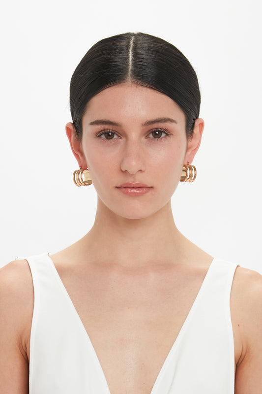 A woman with dark hair pulled back, wearing large Exclusive Frame Hoop Earrings In Gold by Victoria Beckham and a white, sleeveless V-neck top, stares directly into the camera against a plain white background.