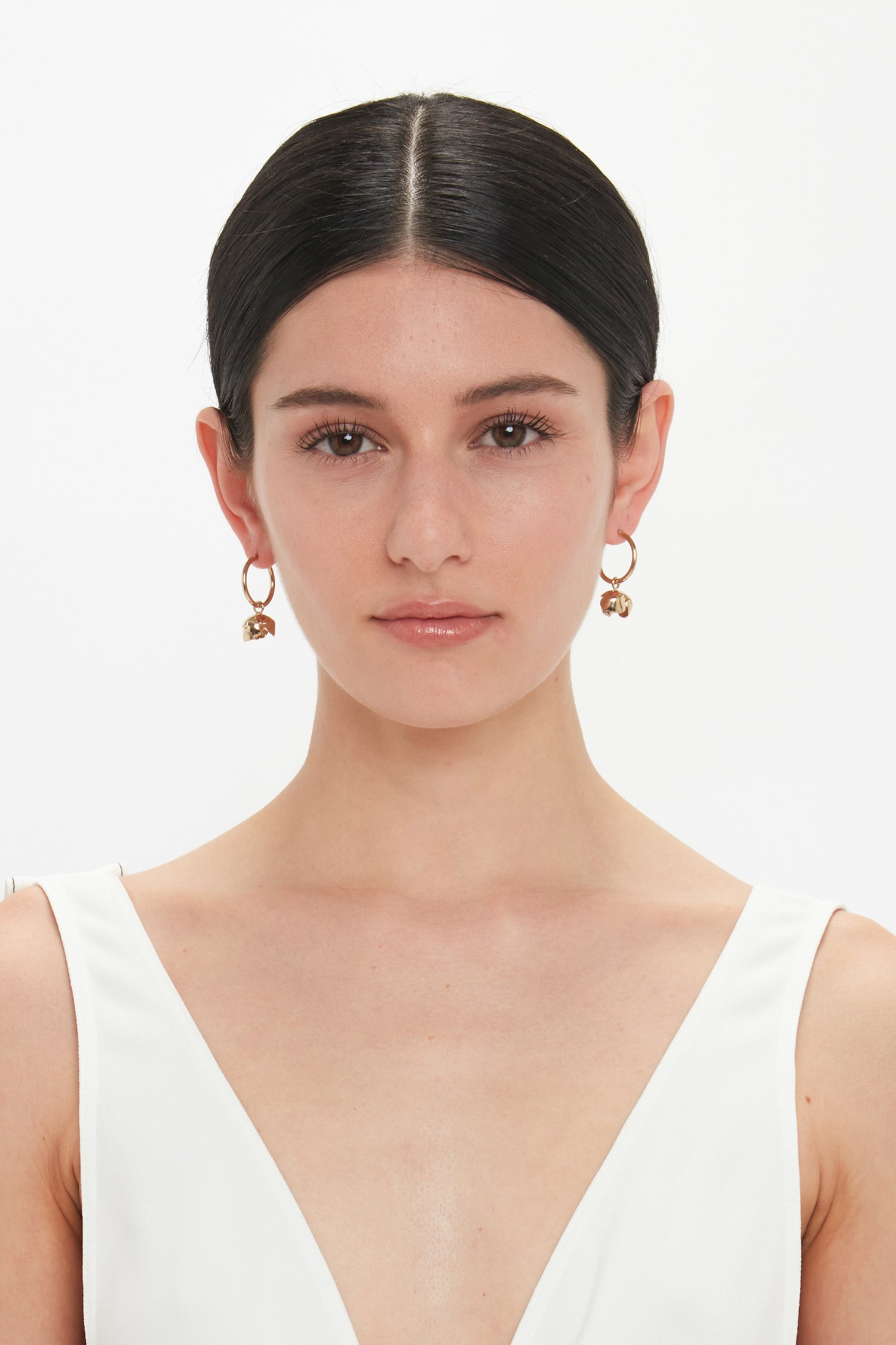 A woman with straight dark hair is wearing a white sleeveless top and Exclusive Camellia Flower Hoop Earrings In Gold by Victoria Beckham. She has a neutral expression and is looking directly at the camera. The intricate earrings are hand-crafted in Italy, adding a touch of elegance to her look.