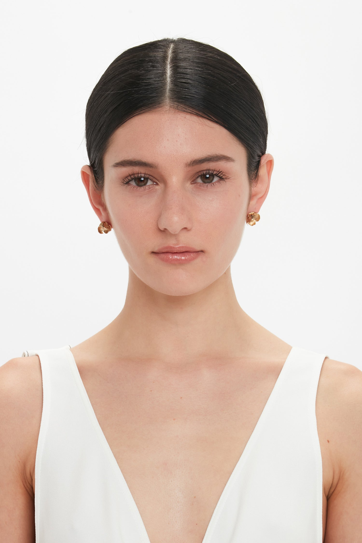 A woman with dark hair parted in the middle wears a white sleeveless top and Exclusive Camellia Flower Stud Earrings In Gold by Victoria Beckham, posing against a white background.