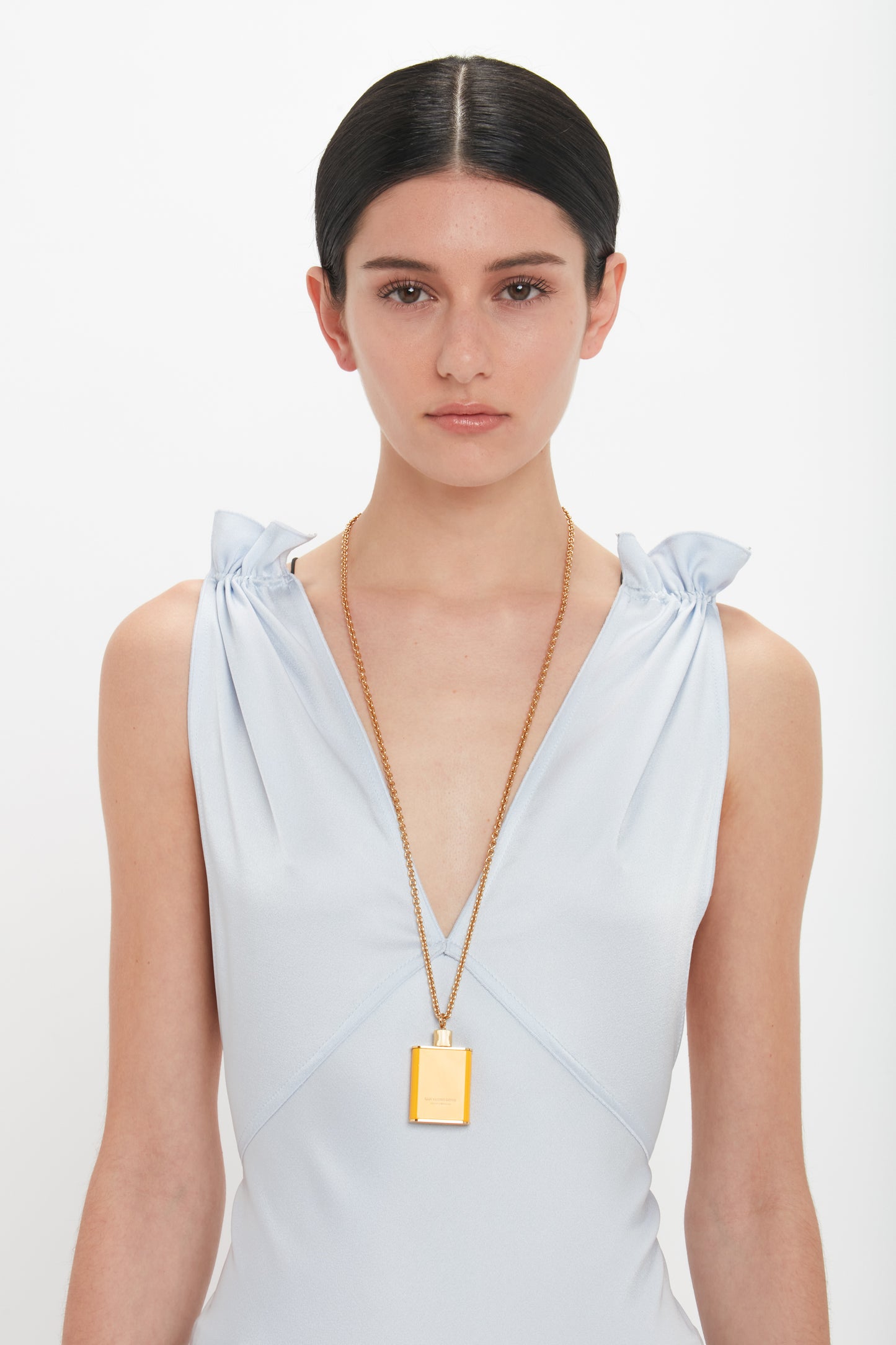 A woman with dark hair, tied back, wearing a light blue sleeveless dress with knotted shoulders. She has a neutral expression and is wearing a long, gold brushed brass chain necklace with a rectangular pendant that complements her unique jewellery style, the Perfume Bottle Necklace In San Ysidro Drive from Victoria Beckham.