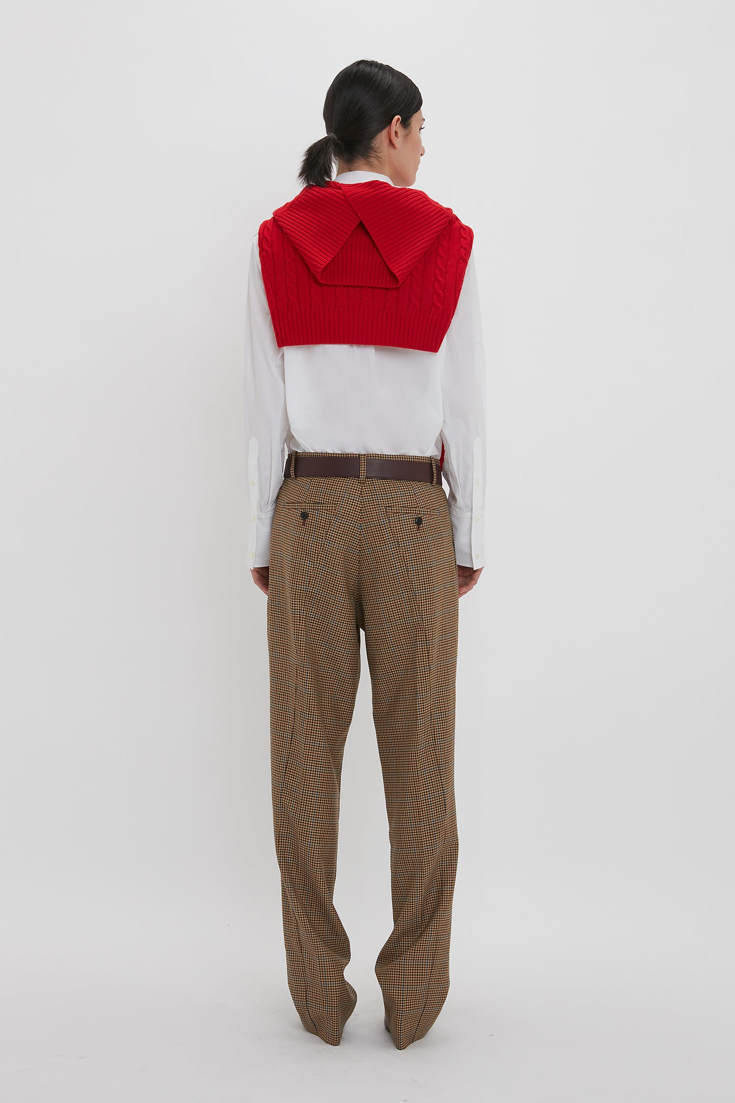 A person stands facing away, donning the Victoria Beckham Asymmetric Chino Trouser In Tobacco with a narrow leg silhouette, a white shirt, and a red knit garment draped over their shoulders.