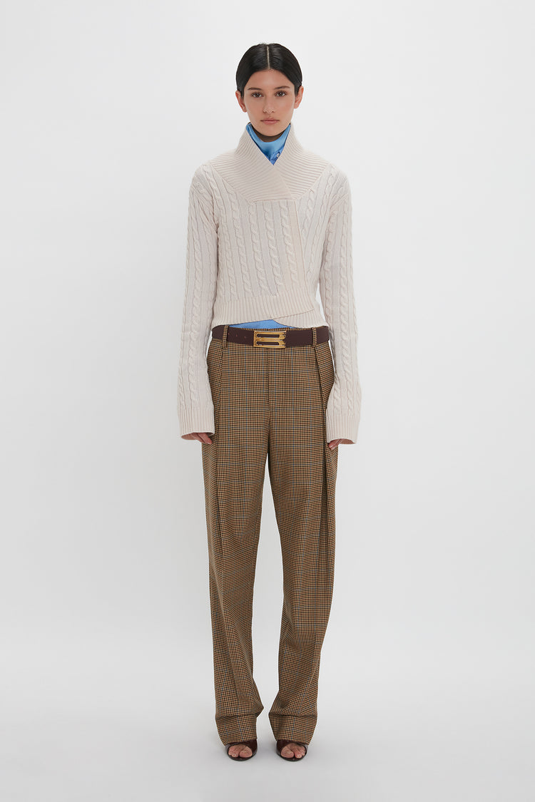 A person stands against a plain background wearing a Victoria Beckham Wrap Detail Jumper In Bone, a blue shirt, brown plaid trousers, and open-toe shoes.