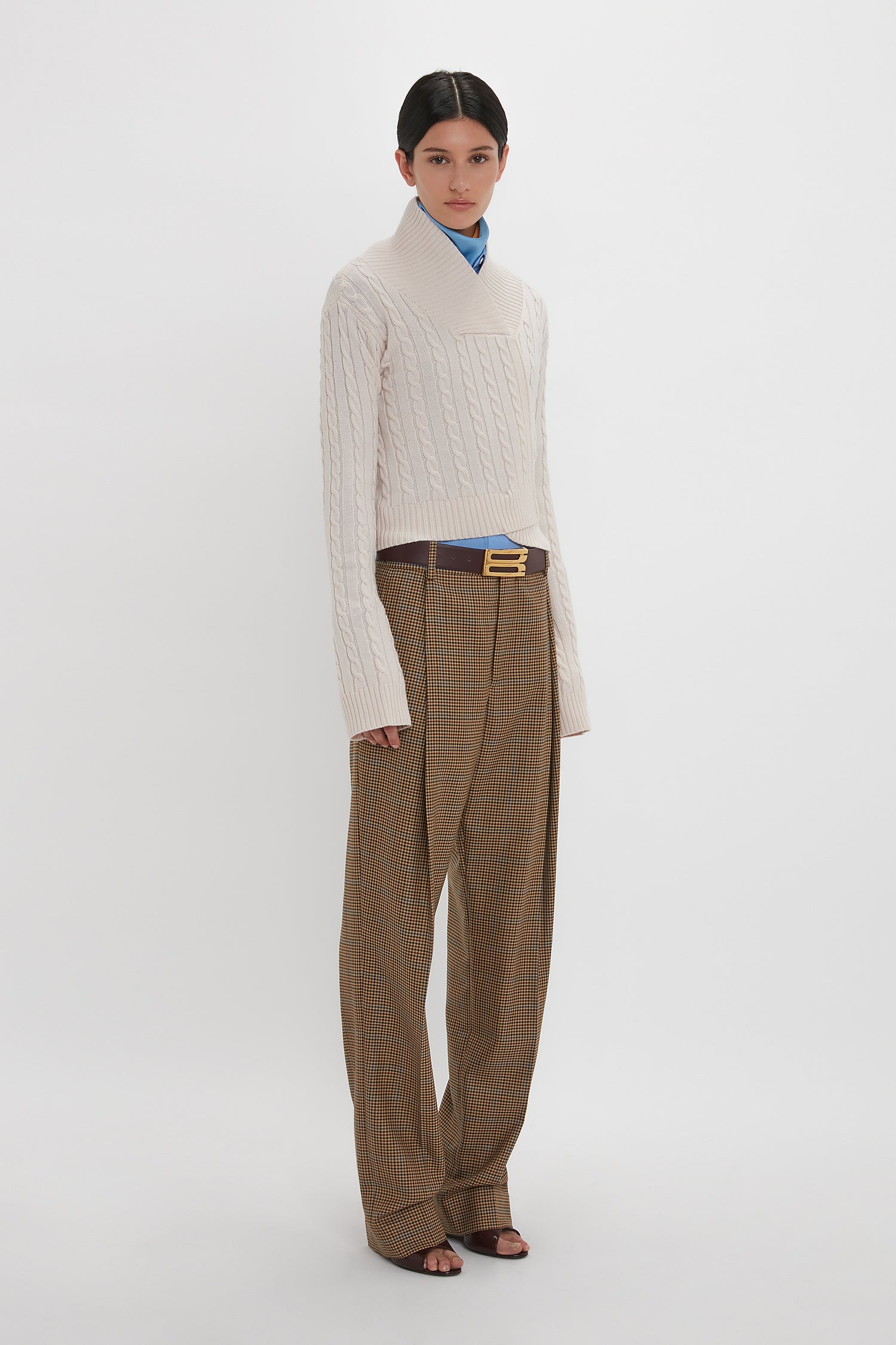 A person stands against a plain background, showcasing a fitted wrap-style silhouette in the *Wrap Detail Jumper In Bone* by *Victoria Beckham*. They pair it with brown checkered trousers, a blue collared shirt, and a brown belt.