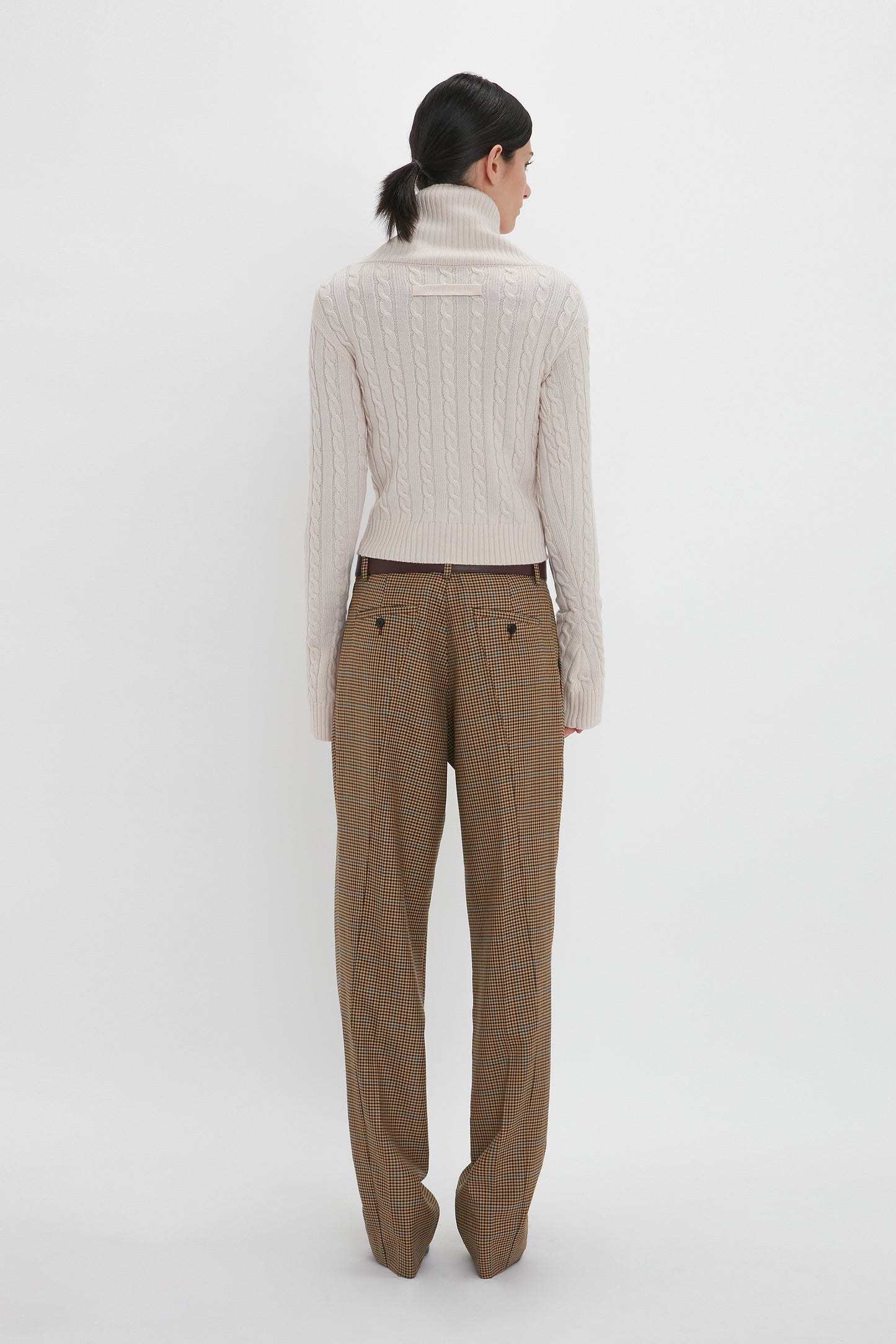 A person stands with their back to the camera, showcasing a Wrap Detail Jumper In Bone by Victoria Beckham, paired with brown plaid trousers.