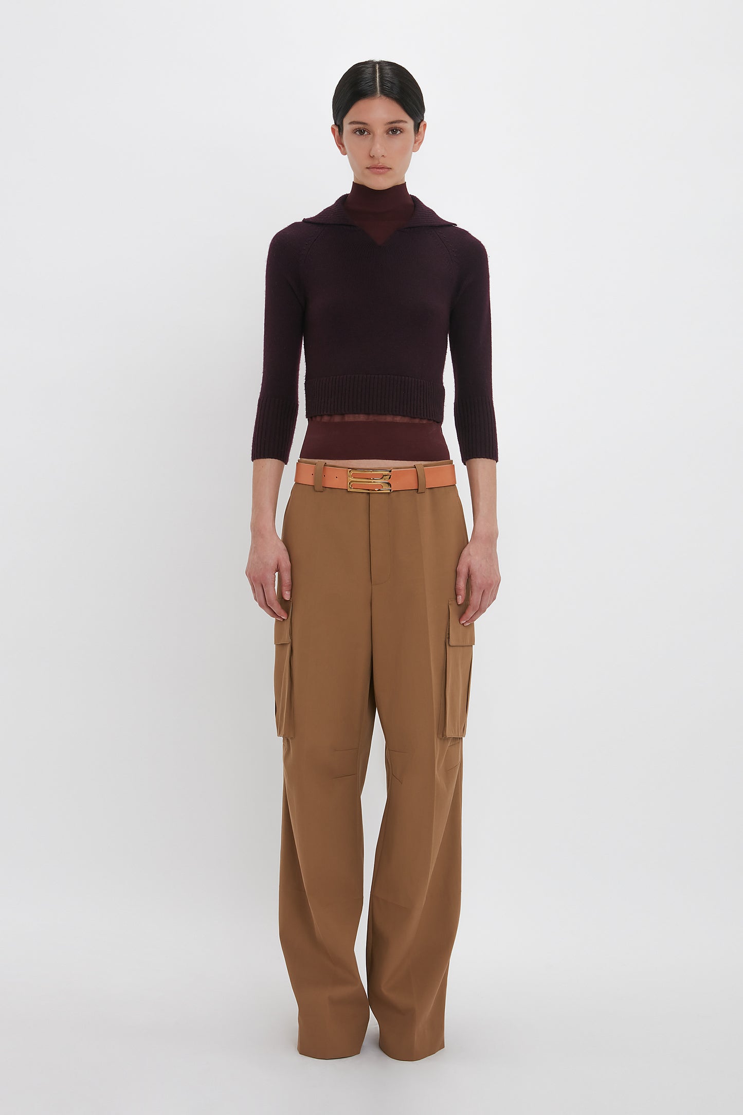 A person stands against a plain white background, wearing the Victoria Beckham Wrap Detail Jumper In Brown, tan high-waisted cargo pants, and a brown belt. They have a neutral expression and their hair is pulled back. The ensemble evokes the sophisticated style of Victoria Beckham.