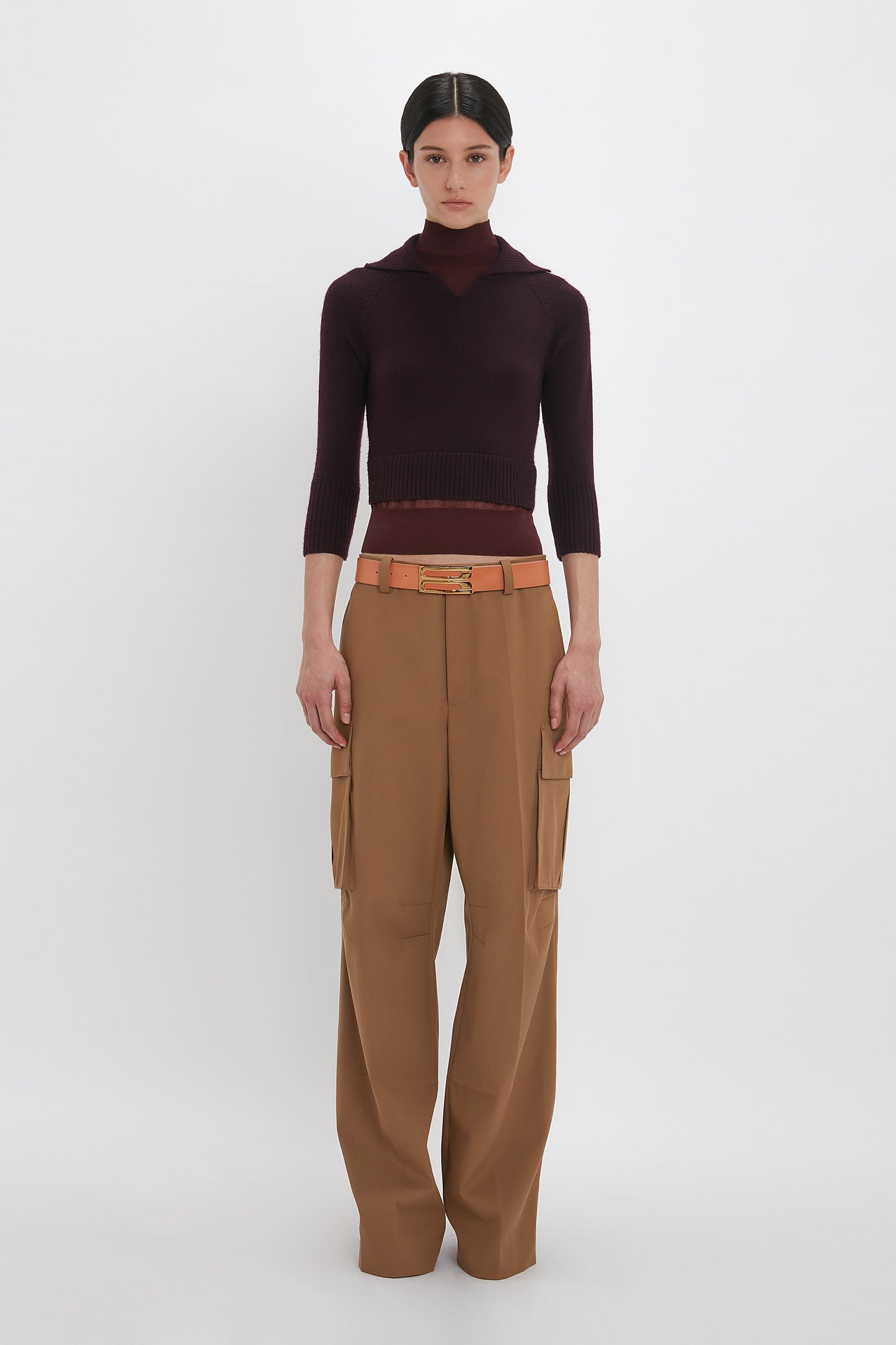 A person stands against a white background, wearing a Victoria Beckham Double Layer Top In Deep Mahogany, brown cargo pants, and a tan belt.