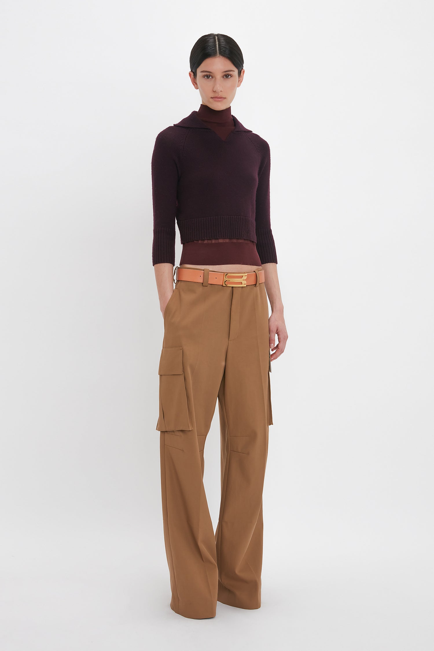 A person stands against a plain white background, wearing a dark maroon Wrap Detail Jumper In Brown by Victoria Beckham and light brown wide-leg cargo pants with a tan belt.