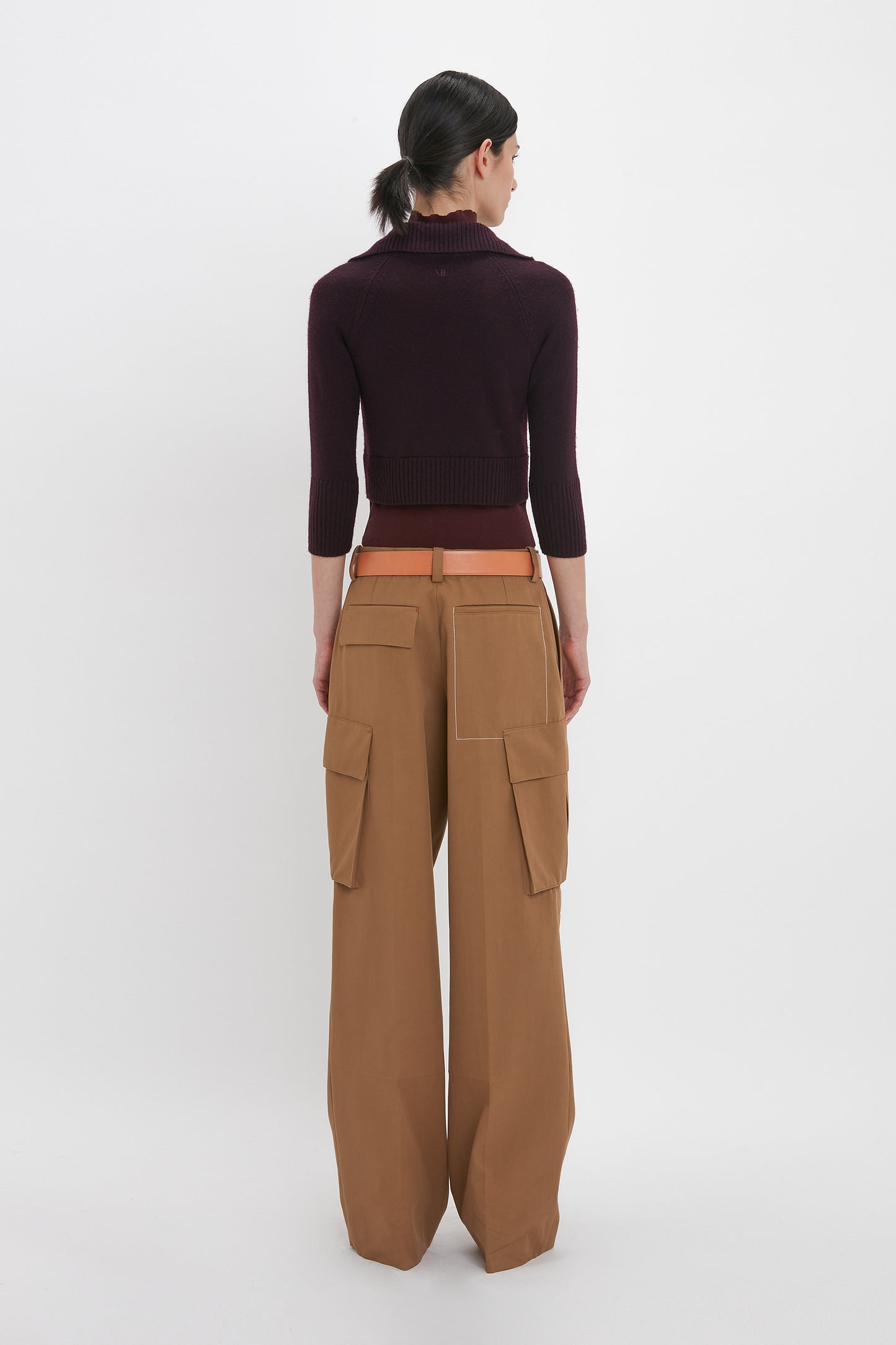 Person standing facing away, wearing a dark luxury knitwear long-sleeve top from Victoria Beckham called the Wrap Detail Jumper In Brown, and high-waisted, wide-leg tan cargo pants with a belt.