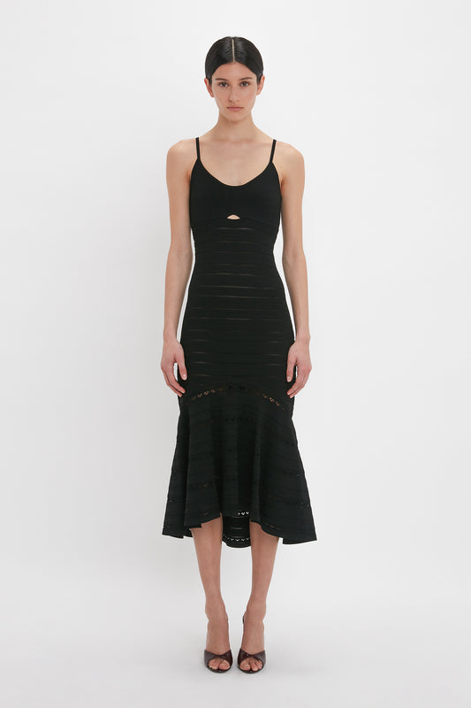 A person with dark hair is wearing a fitted, mid-length black body-sculpting Cut-Out Detail Cami Dress In Black by Victoria Beckham with thin straps and a small cut-out detail in the front, paired with black high-heeled shoes, standing against a plain white background.