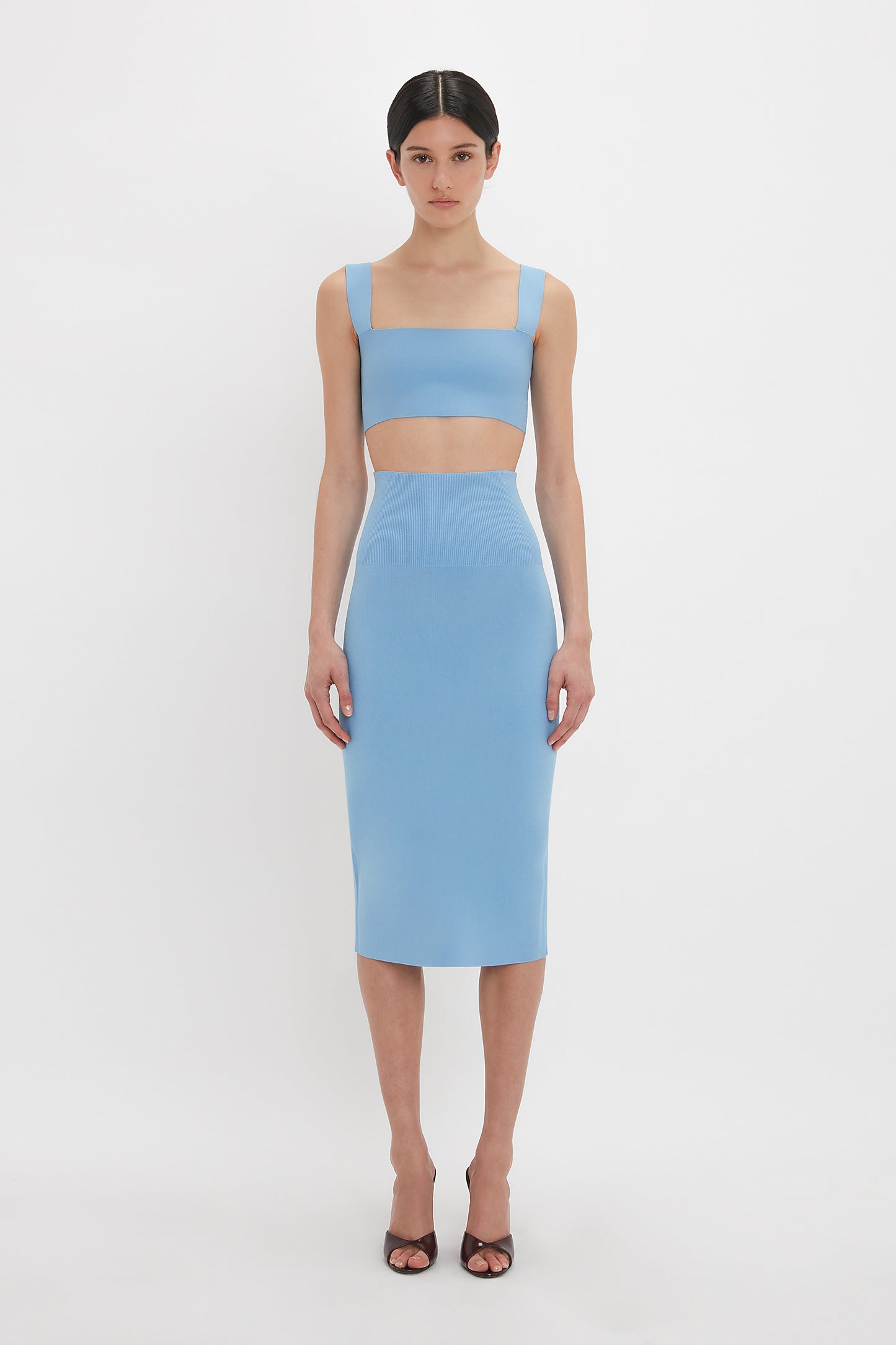 A person stands facing the camera, wearing a light blue, ultra form-fitting two-piece outfit consisting of a Victoria Beckham Strap Bandeau Top In Marina and a fitted midi skirt. They are also wearing brown open-toe high heel sandals.