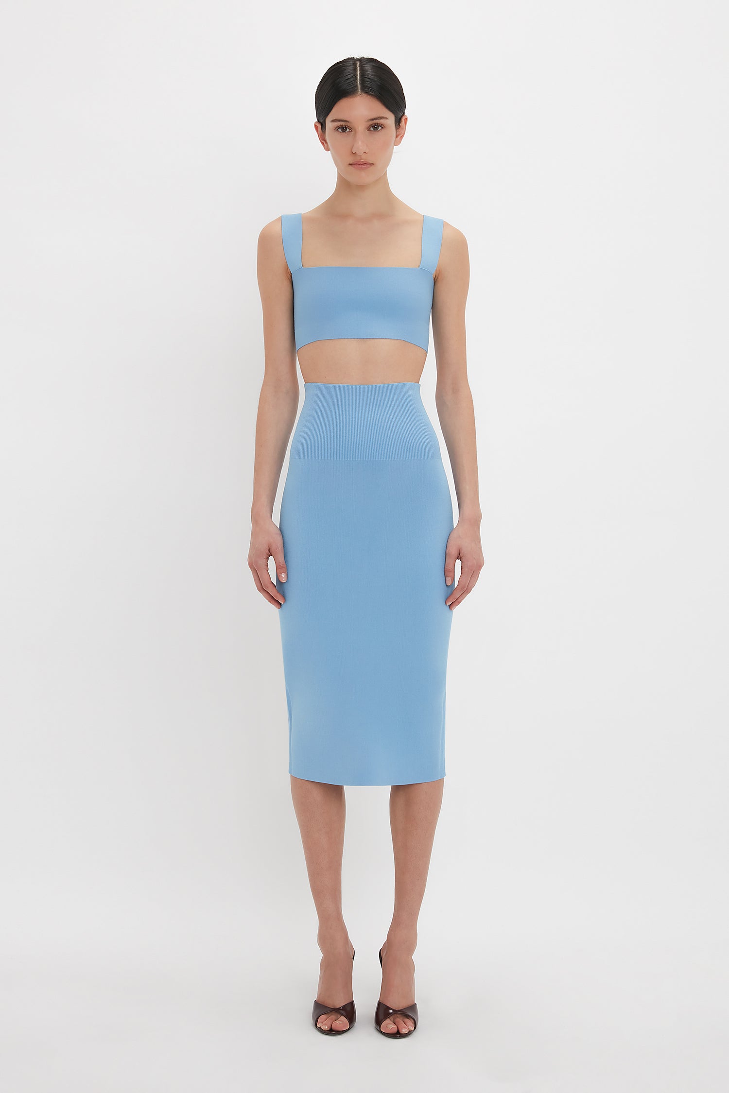 A person is standing against a white background wearing a light blue two-piece outfit consisting of a crop top and a high-waisted, Victoria Beckham Fitted Midi Skirt In Marina, paired with black open-toe heels.