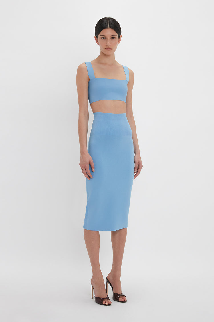 A woman stands against a plain white background, wearing an ultra form-fitting blue Strap Bandeau Top In Marina by Victoria Beckham and matching high-waisted midi skirt with black high-heeled sandals.