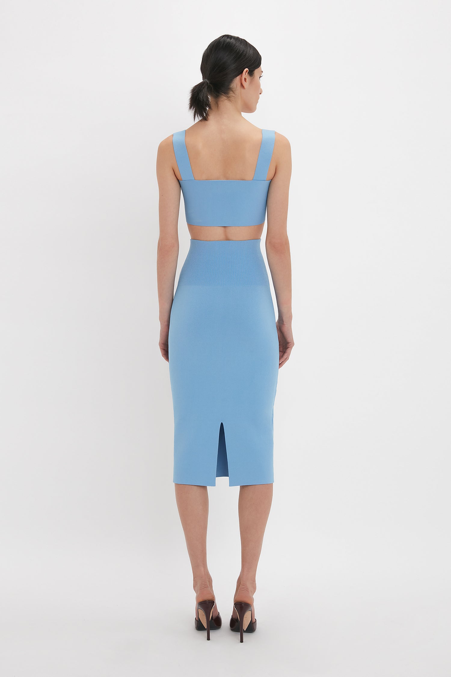 A person stands facing away, wearing a Strap Bandeau Top In Marina by Victoria Beckham with a cutout back and high heels, accentuating an ultra form-fitting silhouette.